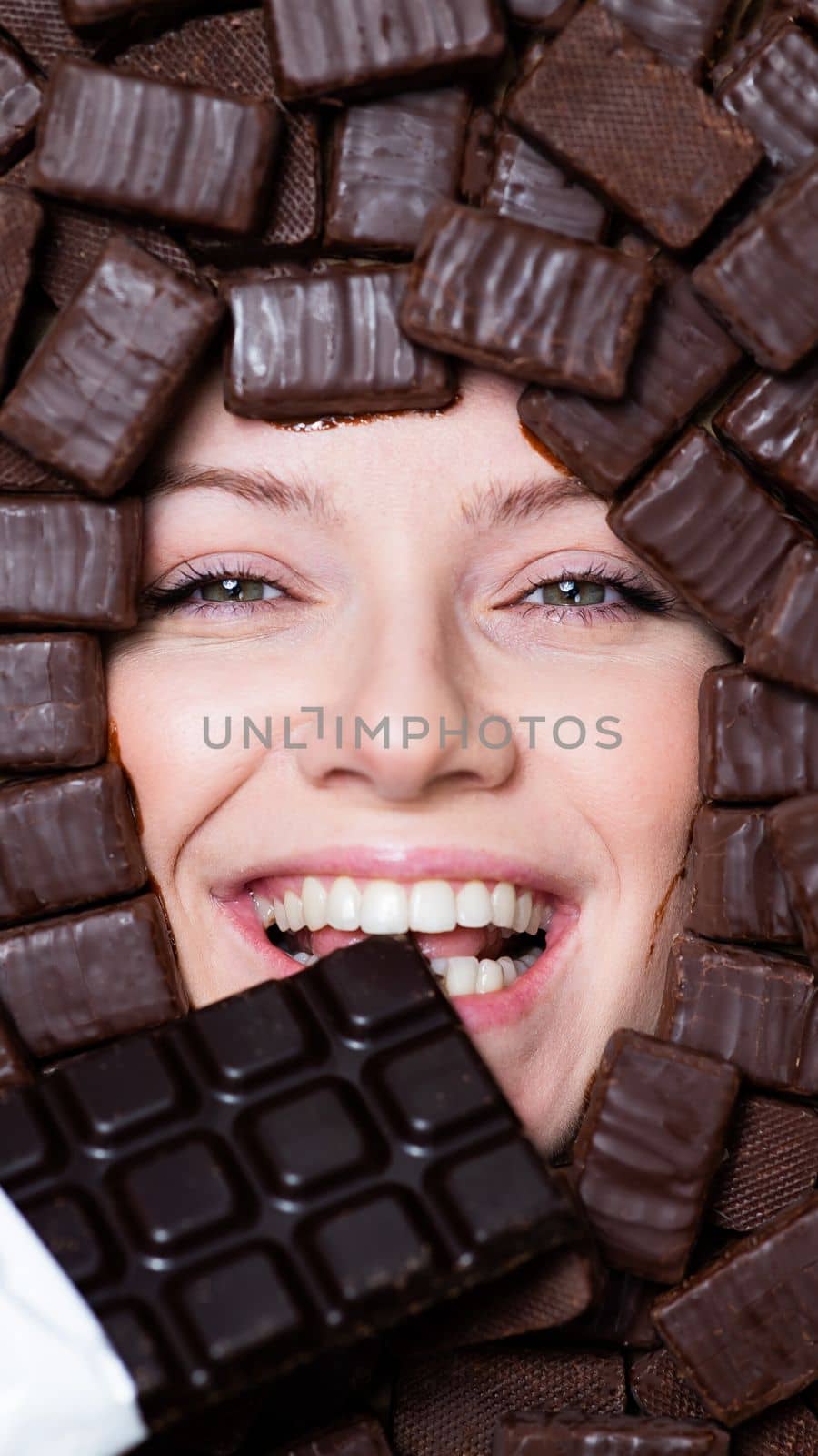 The face of a caucasian woman surrounded by chocolates. The girl eats a bar of chocolate