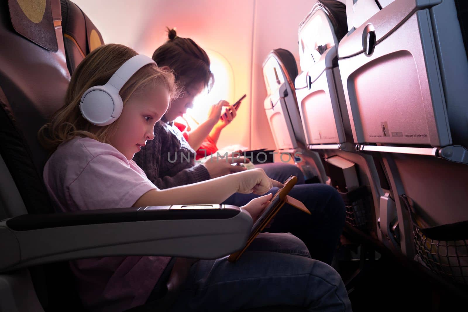 Family journey by plane. Little girl in wireless headphones using digital tablet, boy and woman entertaining with phones