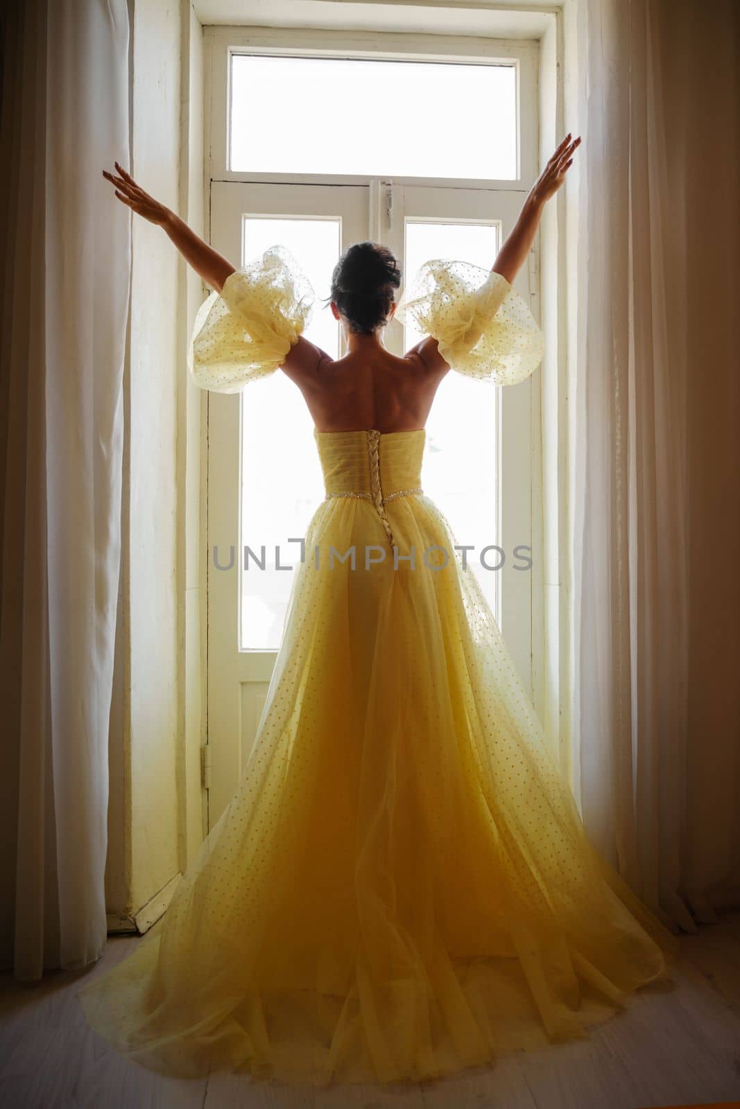A woman's silhouette in a golden luxurious dress against the background of a window holds a curtain with her hands. Elegant lady in a yellow long silk dress with bare back, back view