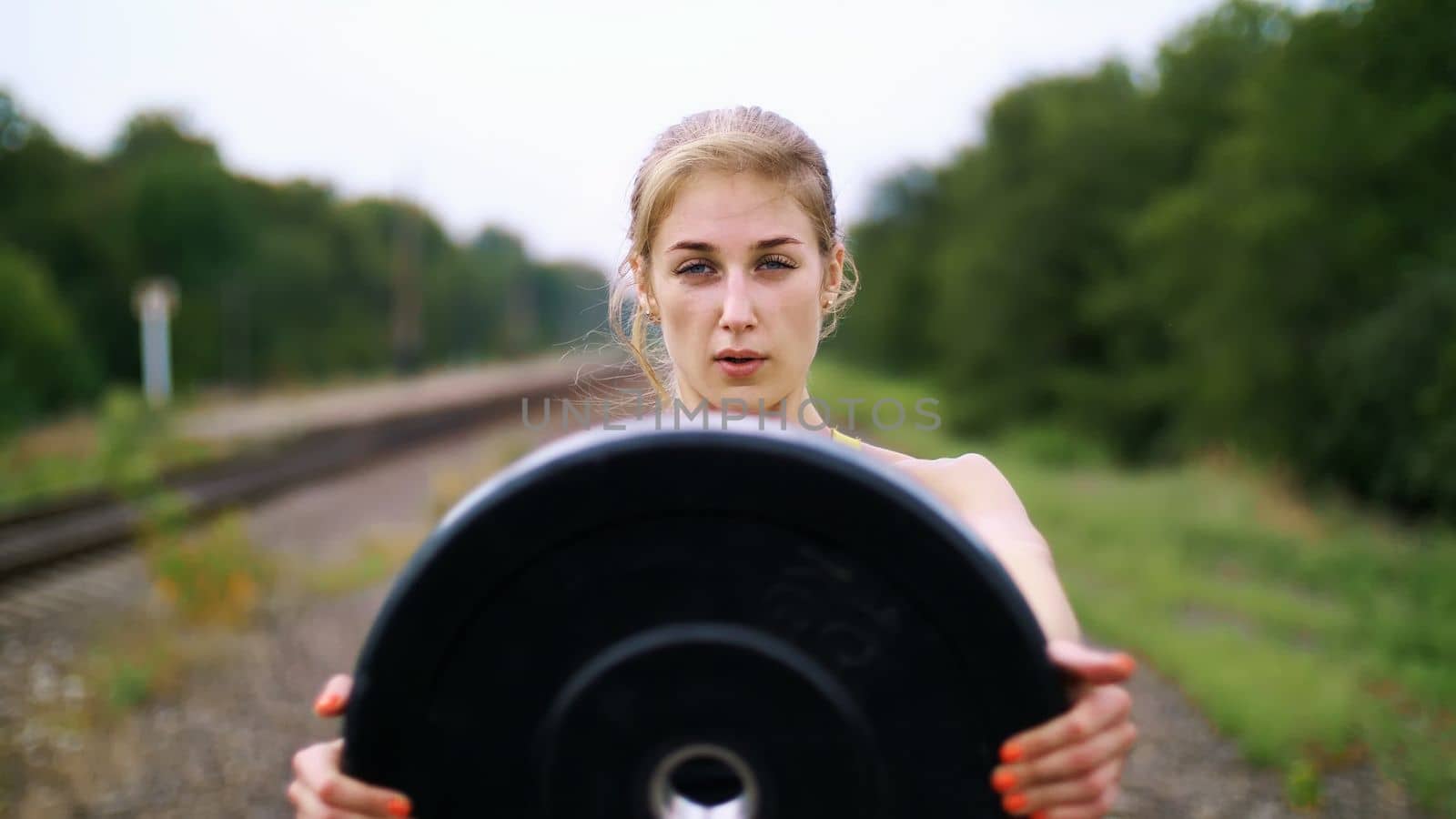 Beautiful sexy athletic young blond woman in black leggings performing exercises with a heavy weight plate, On the railway, on rails, in the summer. Slow motion. portrait. High quality photo