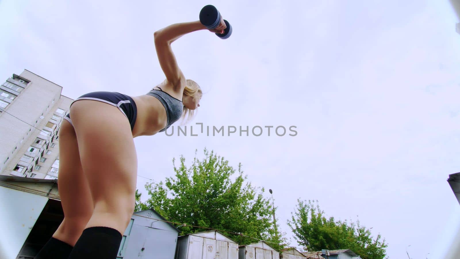Beautiful sexy athletic young blond woman in top, shorts, engaged in crossfit, performs various strength exercises with dumbbells, close-up, near the old abandoned garages. High quality photo