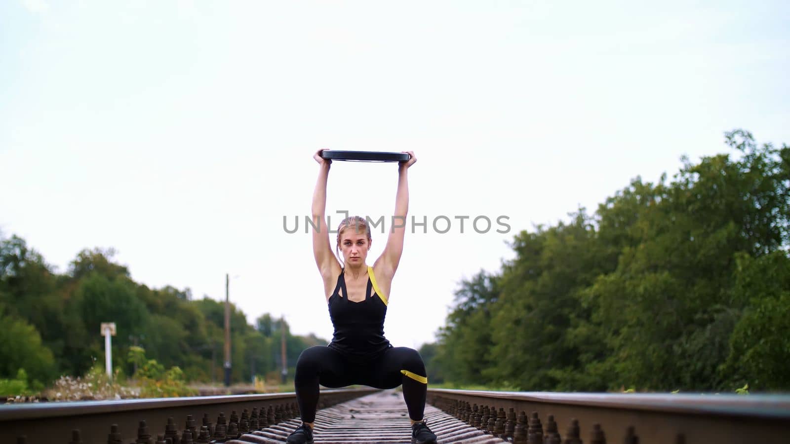 Beautiful sexy athletic young blond woman in black leggings performing exercises with a heavy weight plate, On the railway, on rails, sleepers, in the summer. Slow motion. High quality photo