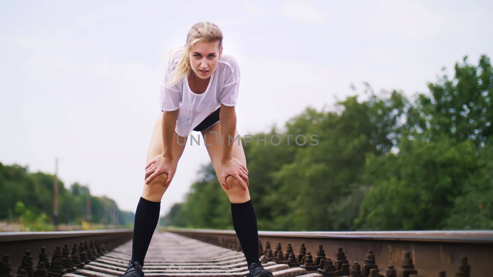 Beautiful sexy athletic young blond woman in top and shorts, running, jogging, Stops, rests, breathes heavily, is tired, looks at the camera. On the railway, in the summer. High quality photo