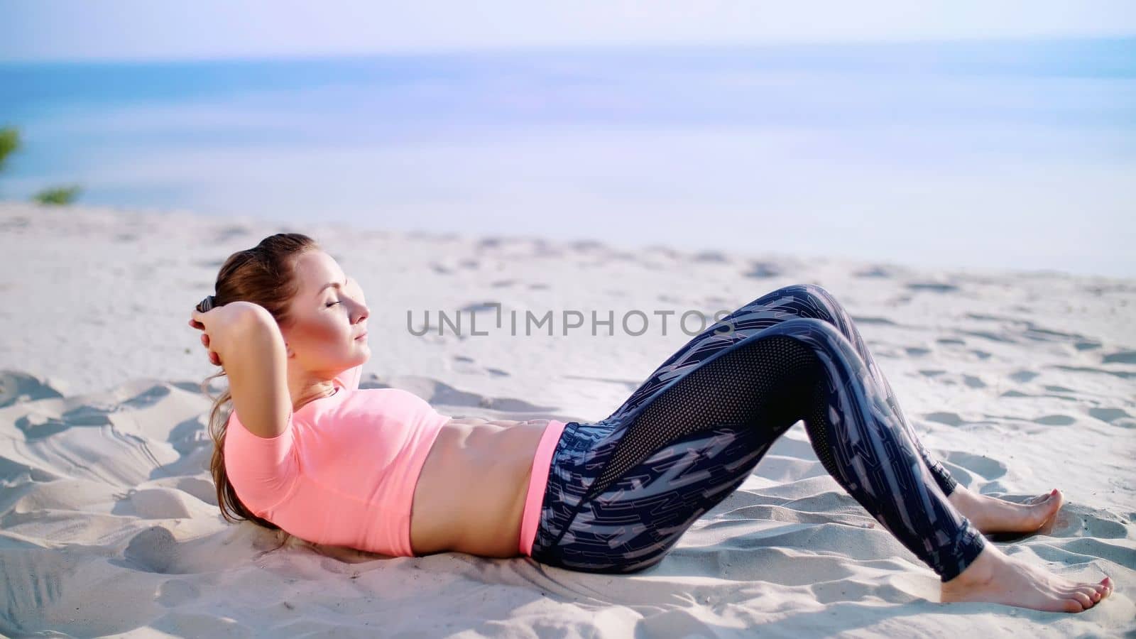 Healthy, young beautiful woman performs exercises for muscles of the press, picks up the body, pumps the press lying on the sand, on the beach, at sunrise. High quality photo