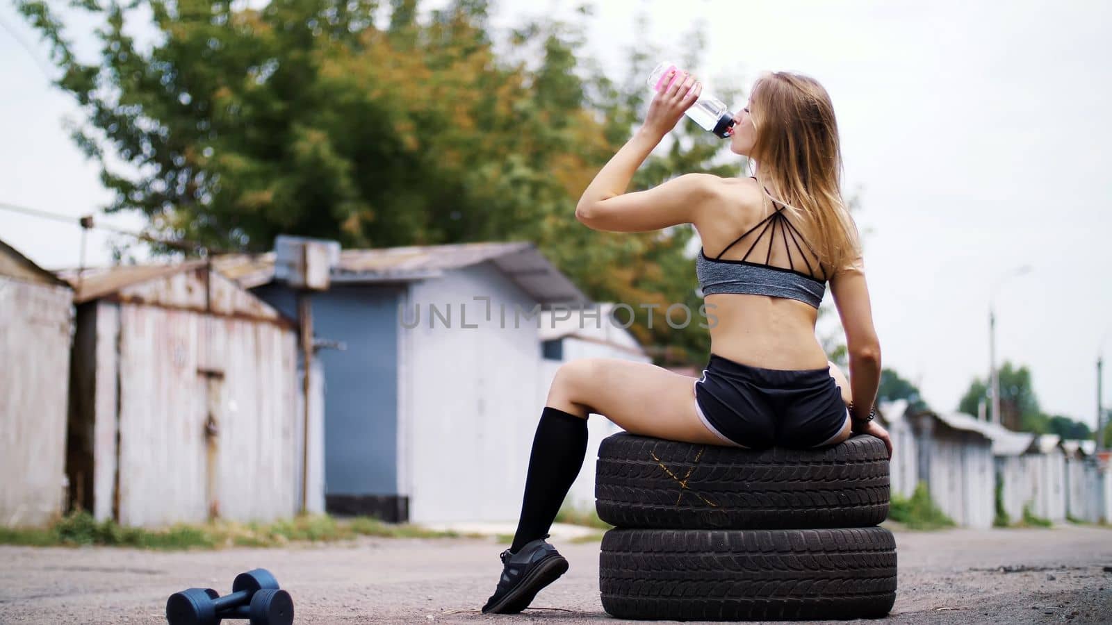 Beautiful sexy athletic young blond woman in top and shorts sitting on tires and drinking water from a bottle, after a crossfit workout. In summer, near the old abandoned garages. High quality photo