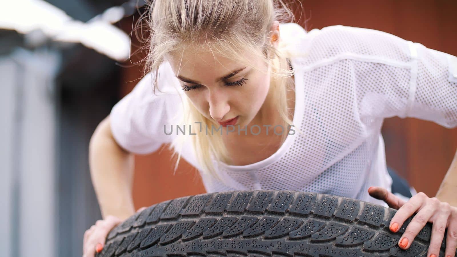 sexy athletic young blond woman in shorts, performs various strength exercises with the help of tires, push-ups, In summer, near old abandoned garages. close up. High quality photo