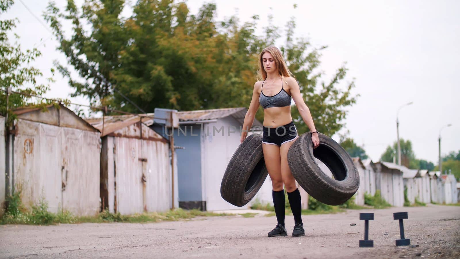 sexy athletic young blond woman in shorts, performs various strength exercises with the help of tires, lifting weights. In summer, near old abandoned garages. High quality photo