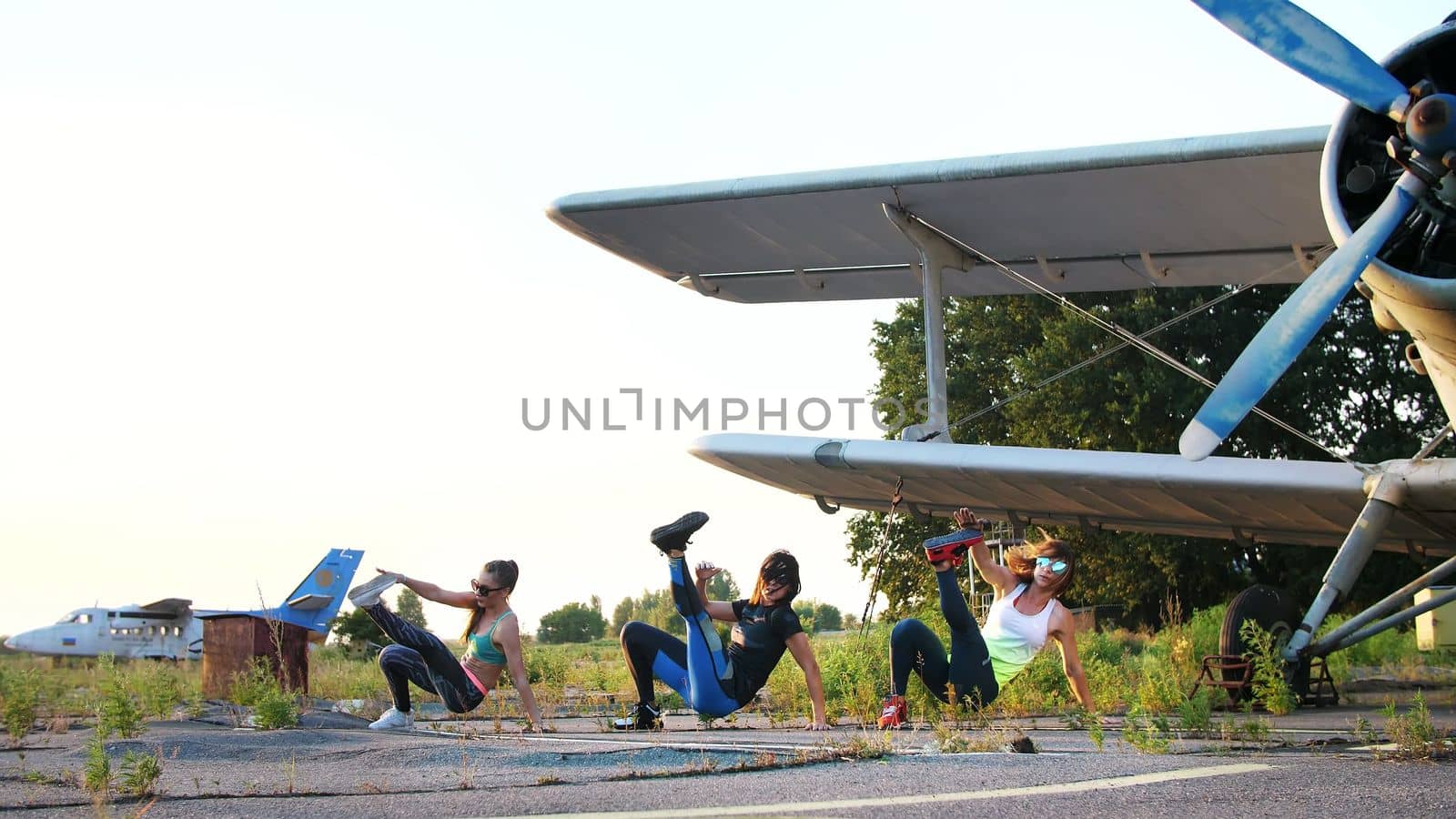 Beautiful, athletic, young women in sunglasses, in tights, perform synchronously different strength exercises, jumps, push-ups, lifting legs.on an abandoned airfield, near plane. High quality photo