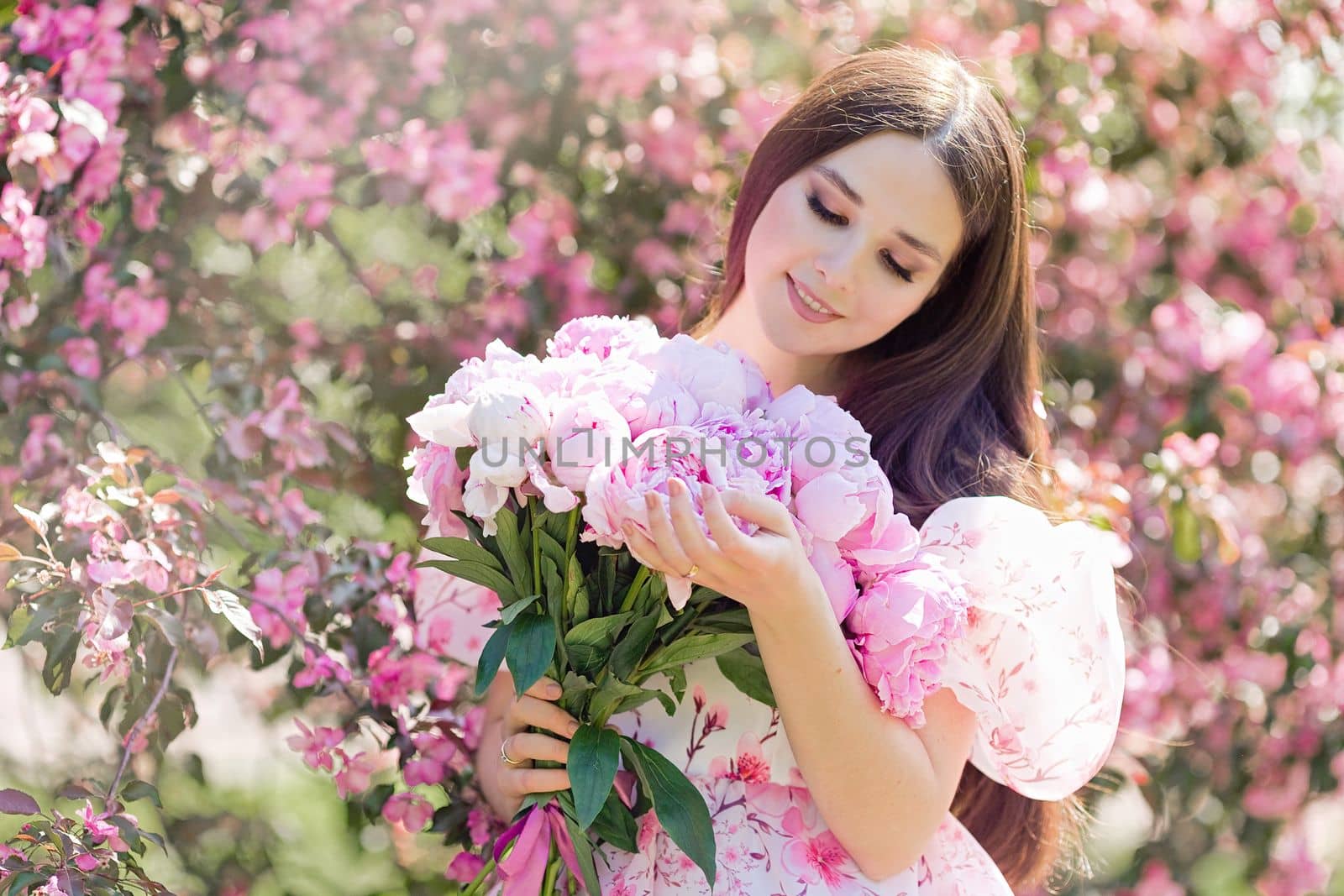 A happy brunette in a light pink dress, with a large bouquet of pink peonies, stands near pink blooming apple trees, in summer in the garden on a sunny day, a space for copying