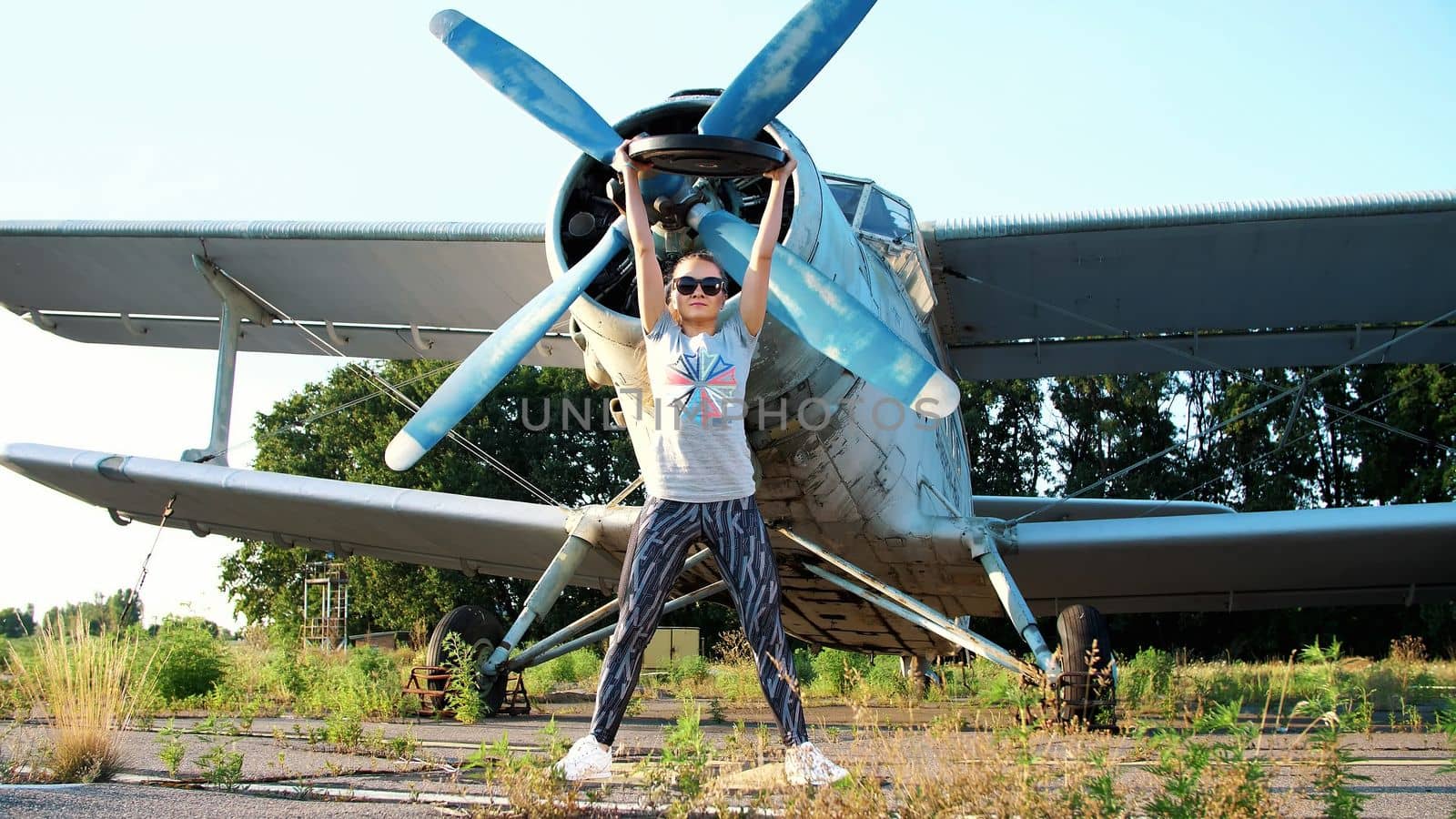 at sunset, athletic, young woman in sunglasses, in tights, doing exercises with weight, heavy weight plate, on an abandoned airfield, against the background of an old plane,. High quality photo