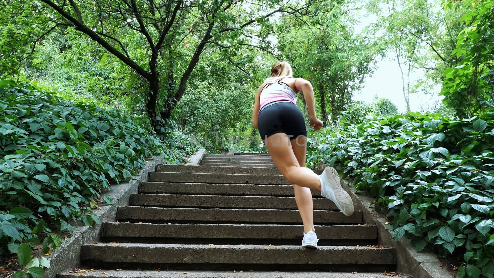 Beautiful sexy athletic young blond woman runs upstairs, in the forest, park, in summer day. Performs exercises to strengthen the different muscles of the legs on the stairs. High quality photo