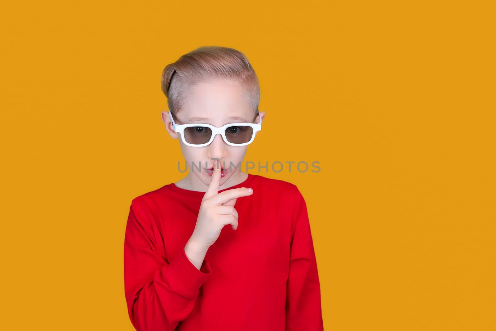 a cheerful child in children's 3D glasses puts his index finger to his lips and makes a quiet gesture