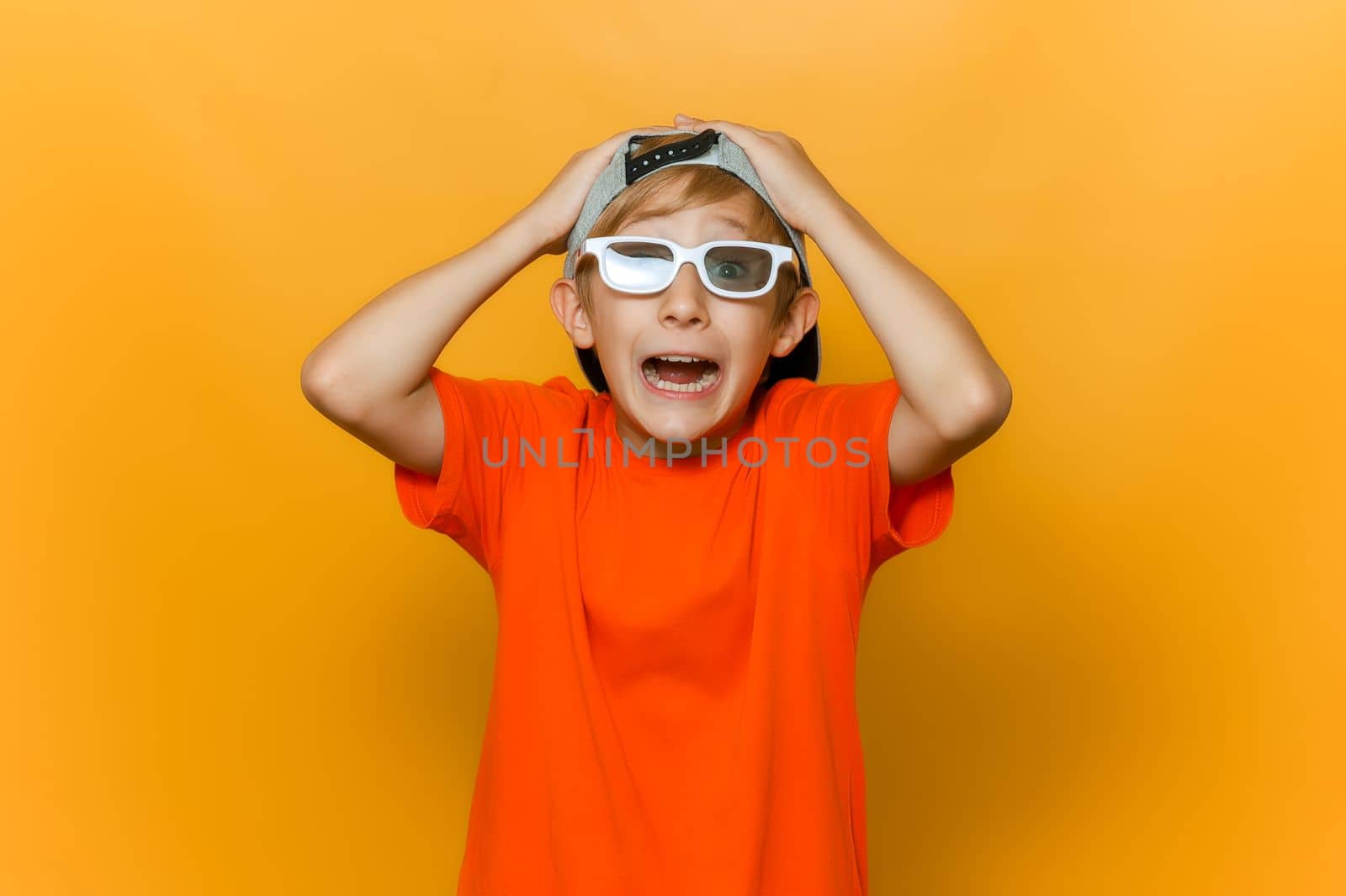 a child in a cap and glasses for watching movies grabbed his head with both hands and shows emotions of fear