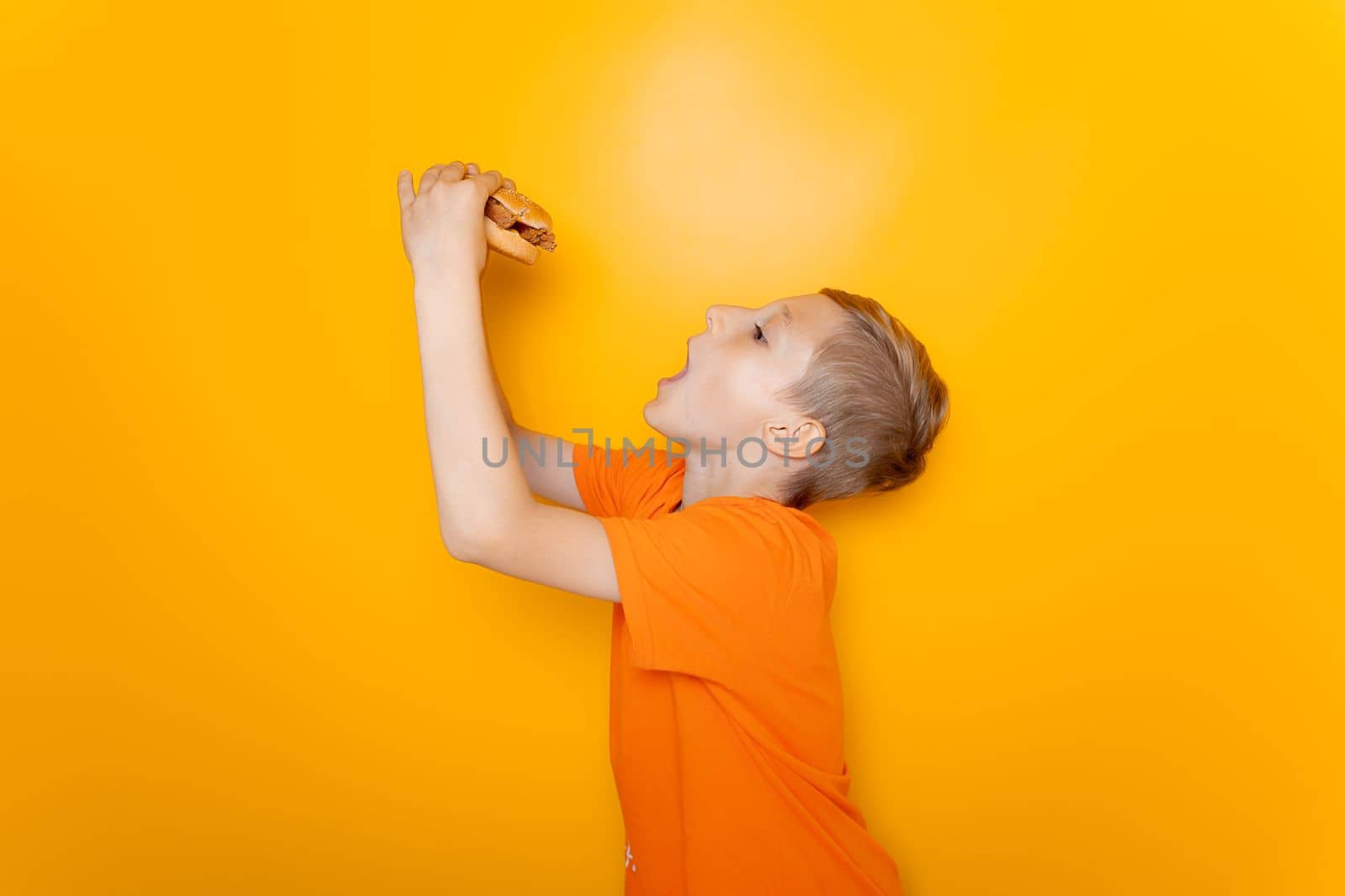 a boy in an orange T-shirt stands sideways and holds a hamburger he wants to eat