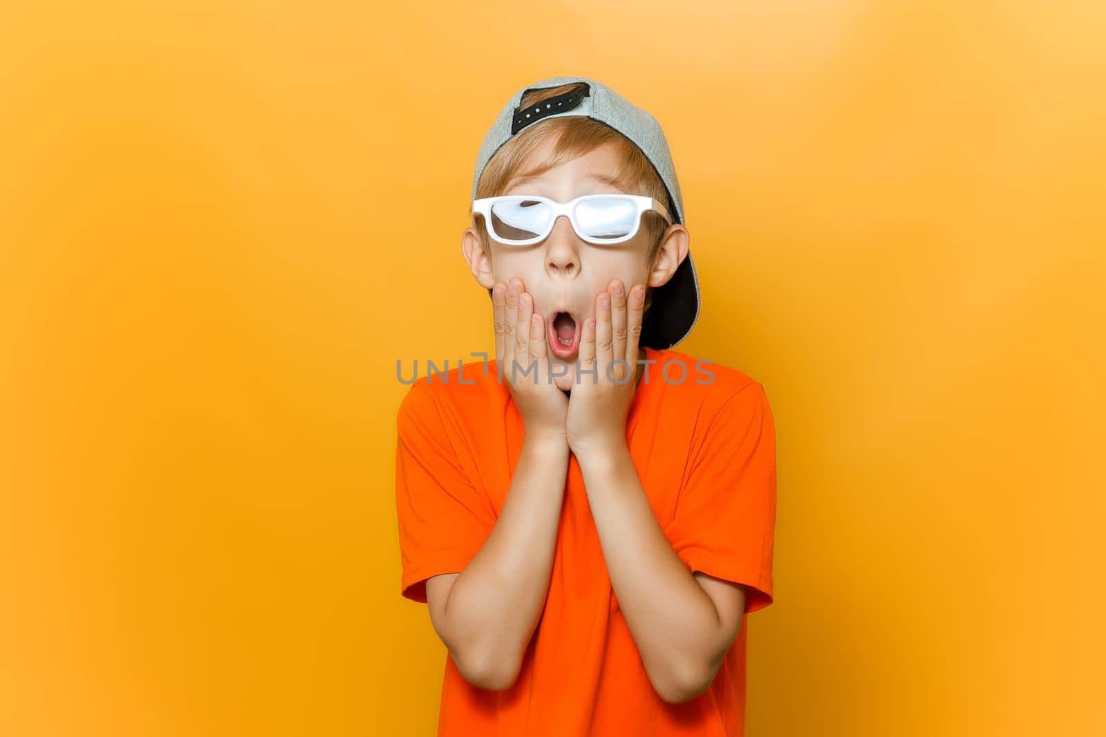 a child in glasses for watching movies opened his mouth in surprise and holds his cheeks with his hands