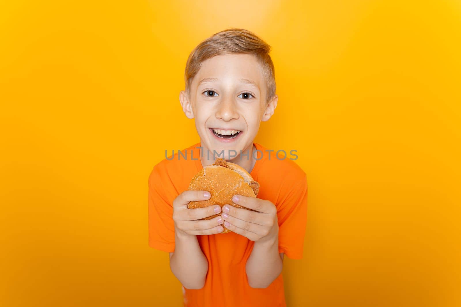 a boy in an orange T-shirt holds a hamburger in front of him and laughs loudly against a yellow background