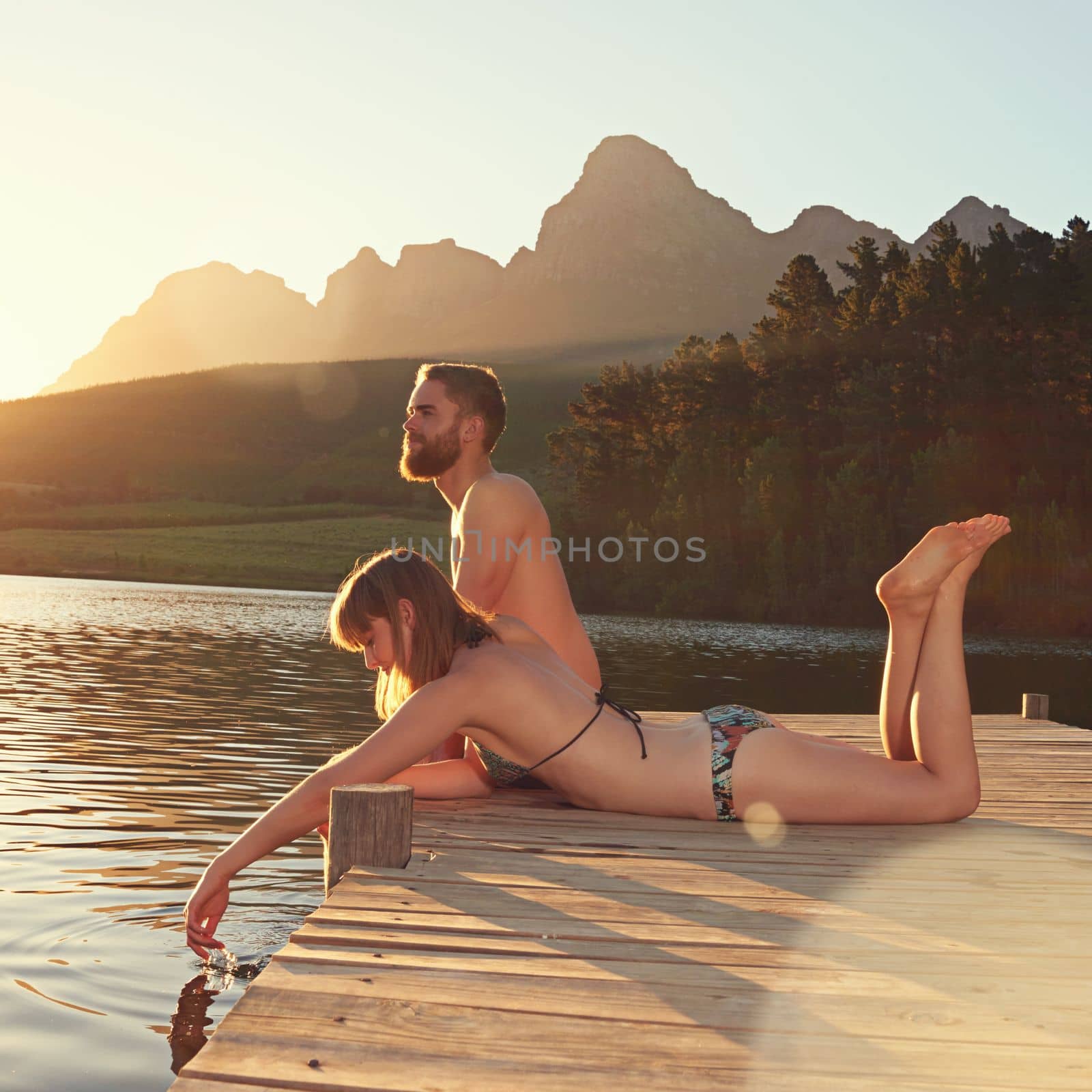 The water is perfect...an affectionate young couple in swimsuits sitting on a dock at sunset. by YuriArcurs