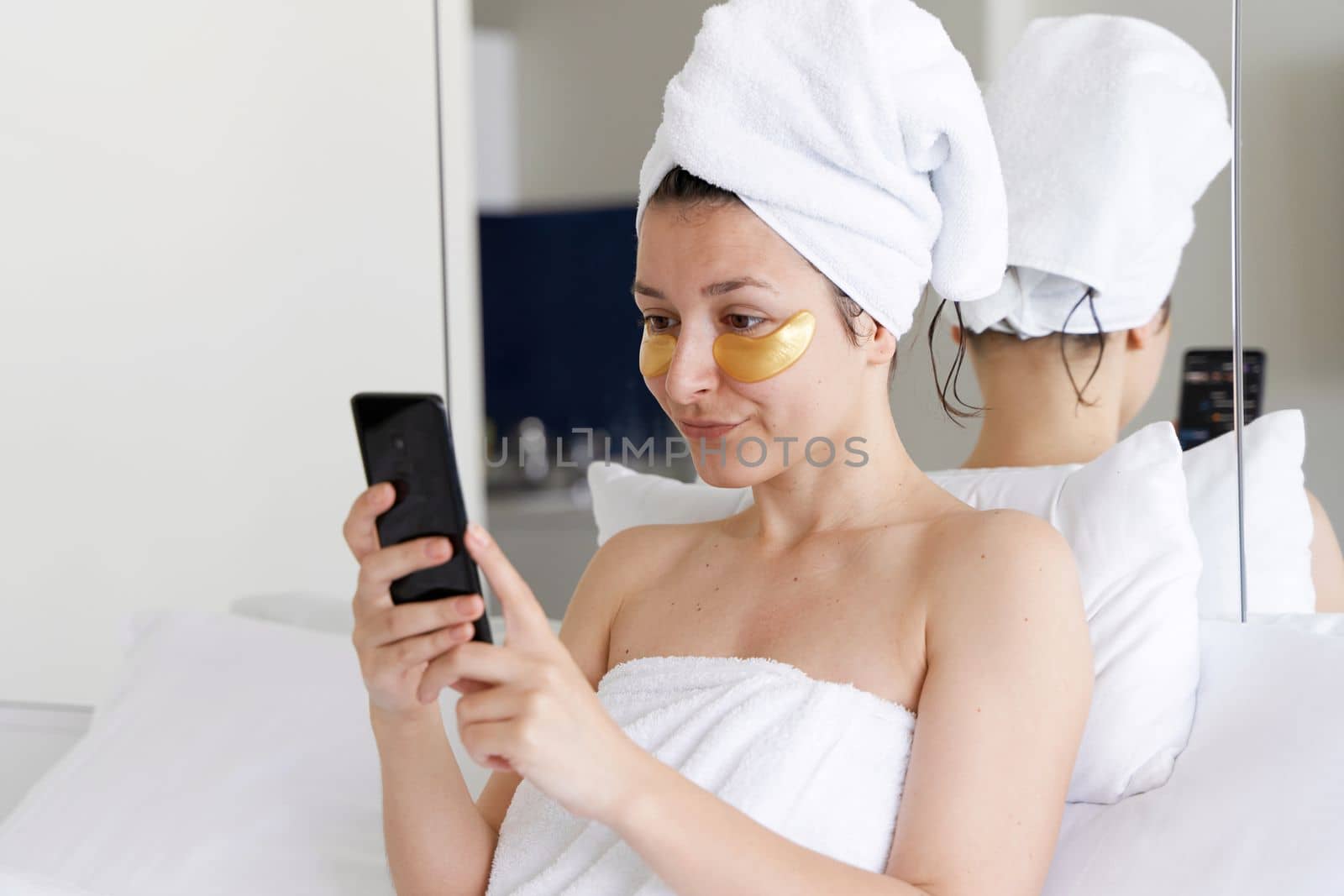 After a shower, the girl is wrapped in a towel and wears cosmetic patches for the skin under the eyes. Looks at the cell phone. Cosmetic procedures at home. Getting ready for a date by Try_my_best