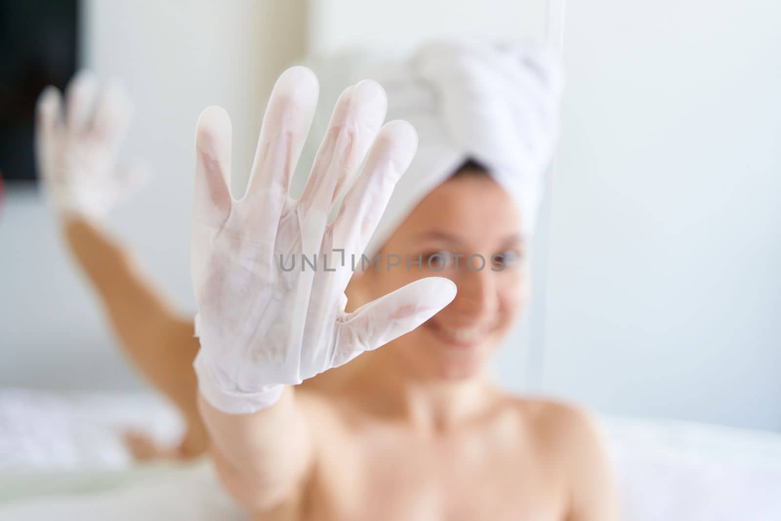 After a shower, a girl wrapped in a towel uses cosmetic gloves to moisturize the skin of her hands. Cosmetic trends for body care at home.