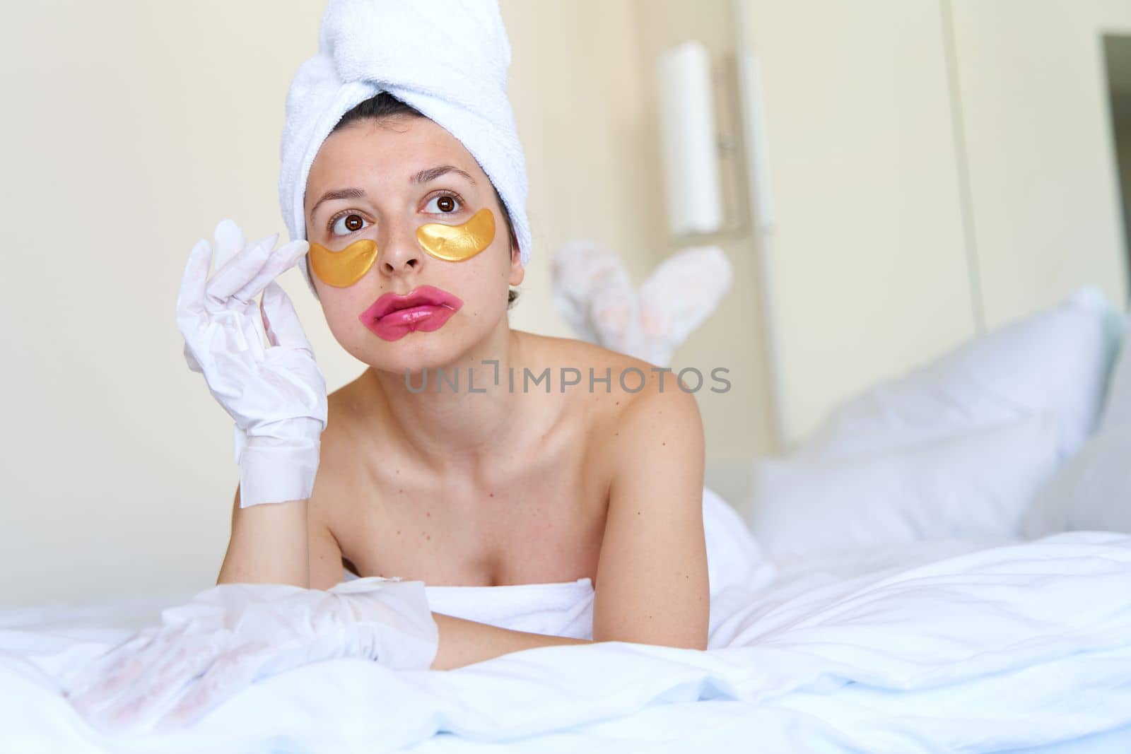 After a shower, a girl wrapped in a towel uses cosmetic patches for the skin under the eyes, lips and gloves to moisturize her hands and feet. Cosmetic trends for body care at home.