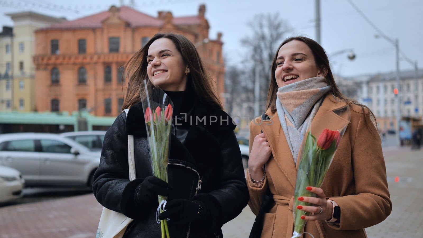 Two joyful friends in a jacket and coat are walking around the city with tulips. by DovidPro