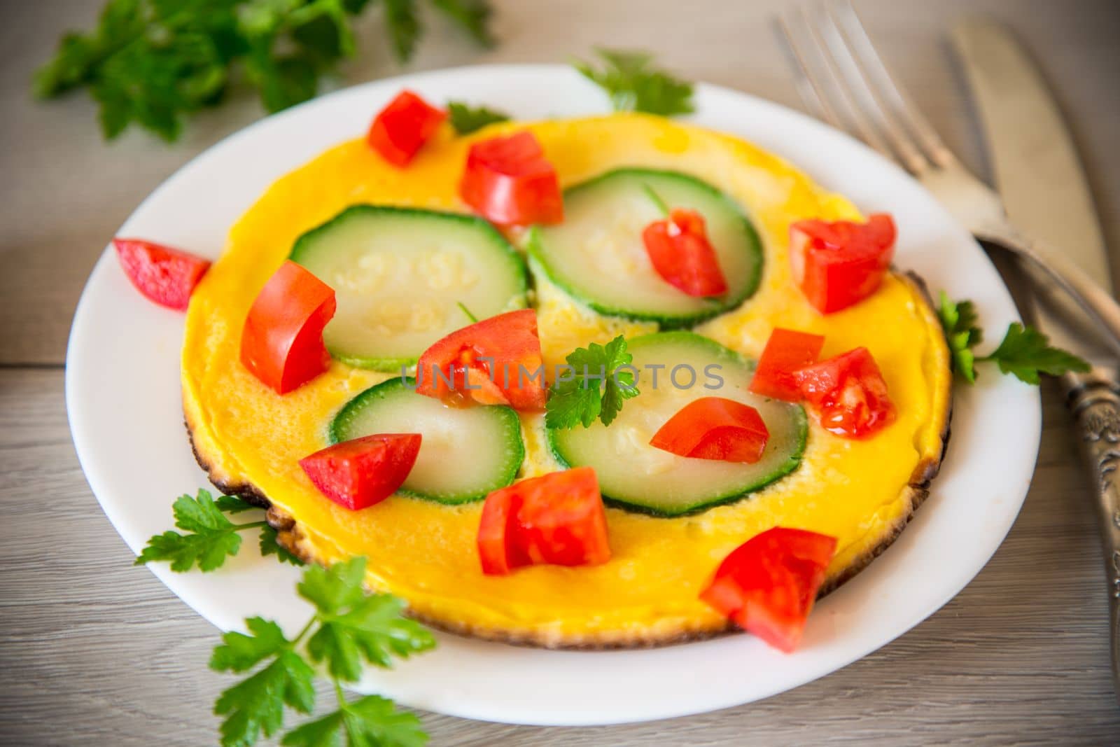 Fried omelet with zucchini, tomatoes, herbs in a plate on a wooden table.