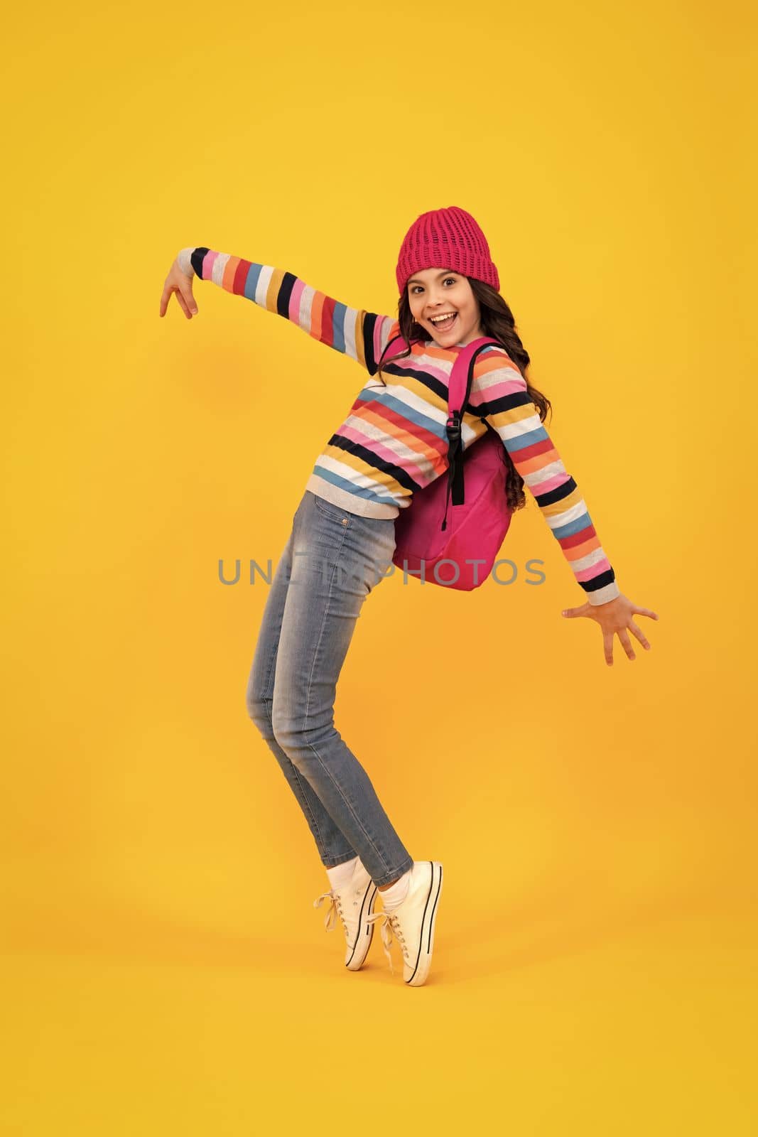 School children in winter hat and sweater with school bag on isolated yellow studio background. Children learning and education. Happy teenager, positive and smiling emotions of teen schoolgirl