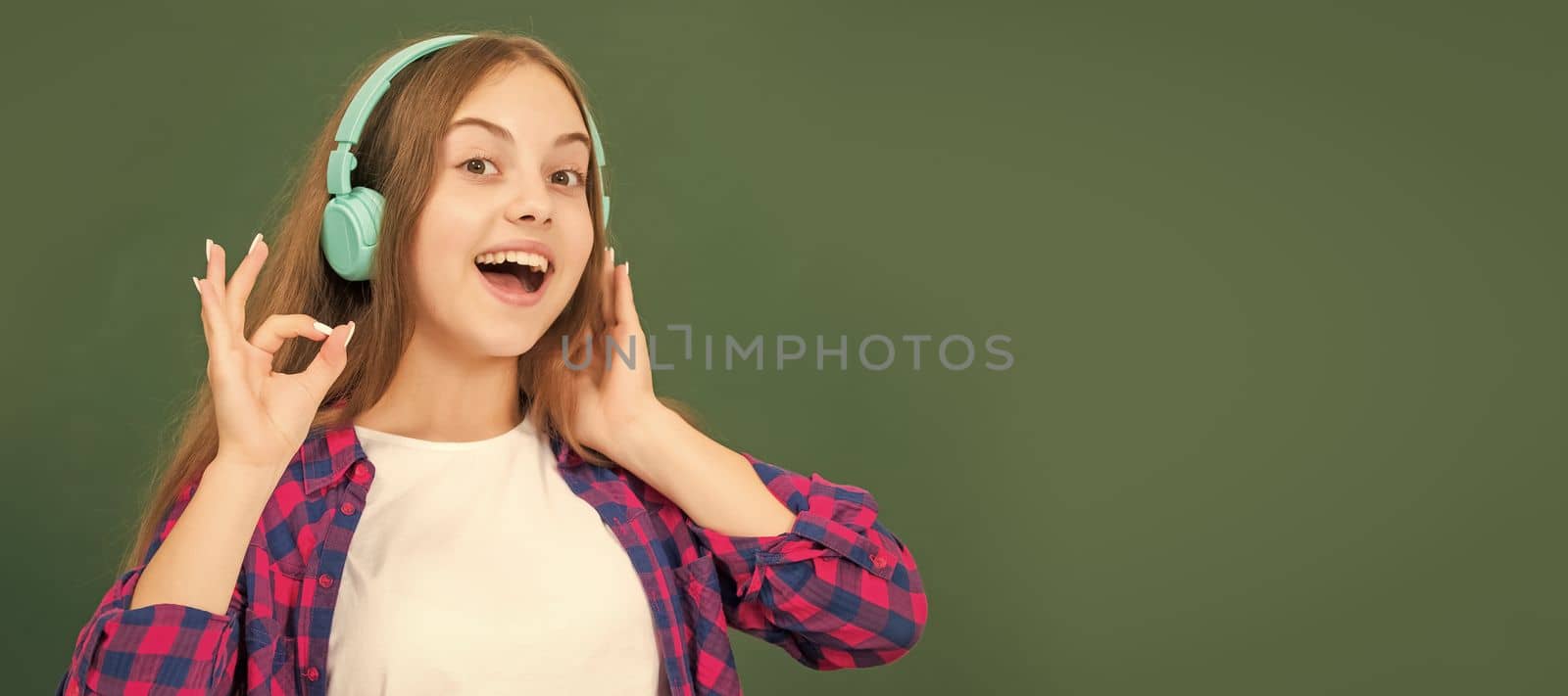 cheerful child listen music in headphones at blackboard. Portrait of schoolgirl student, studio banner header. School child face, copyspace. by RedFoxStudio