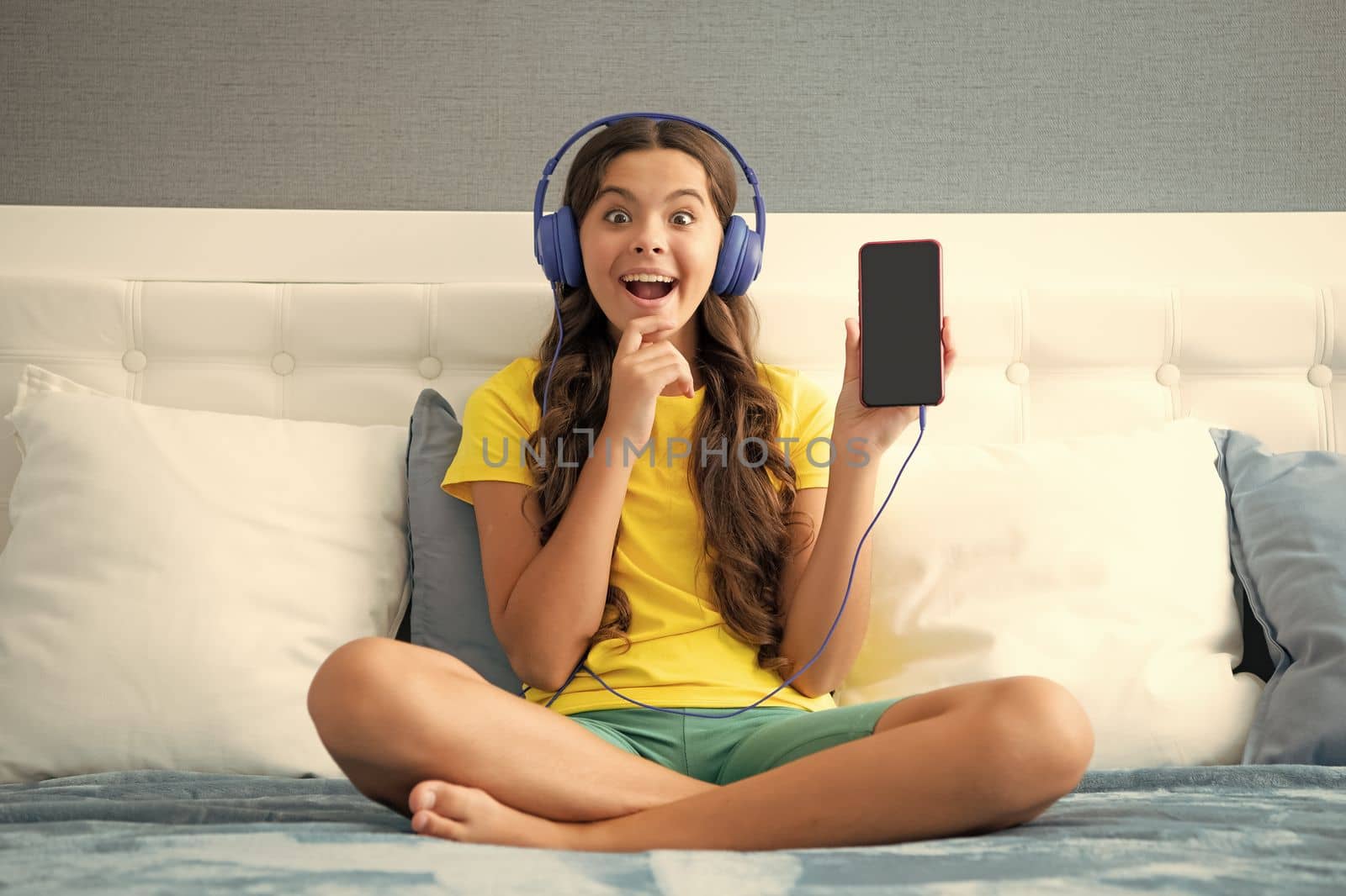 Expressive emotional excited teen girl. Teenager girl in headphones relax on bed at home using phone. Child in earphones showing screen phone