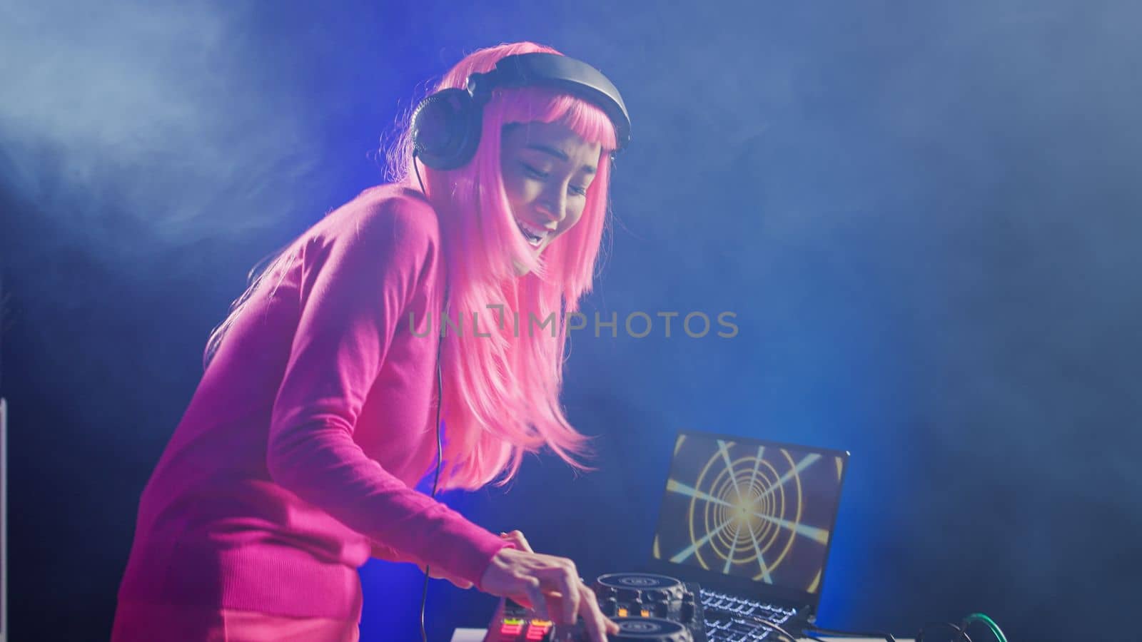 Smiling artist standing at dj table playing electronic music at professional mixer console in studio over pink background. Asian musician performing techno sound, having fun in club at night time