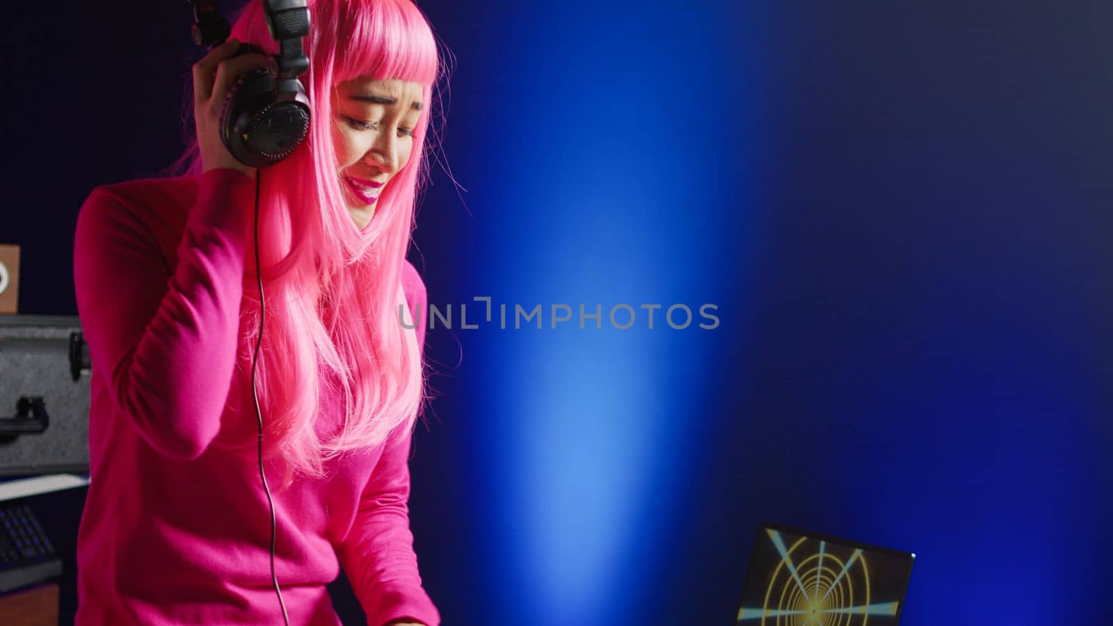Dj with pink hair playing eletronic song at professional mixer console, listening music into headset. Asian perfomer enjoying mixing sounds, dancing with fans during night time in club.