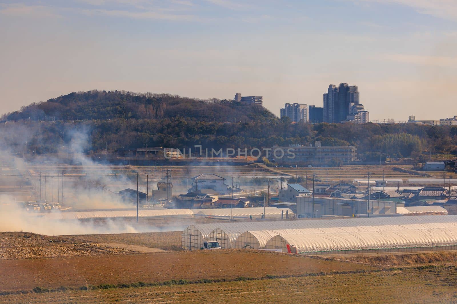 Smoke from fire rises over greenhouses and fields by houses in rural area. High quality 4k footage