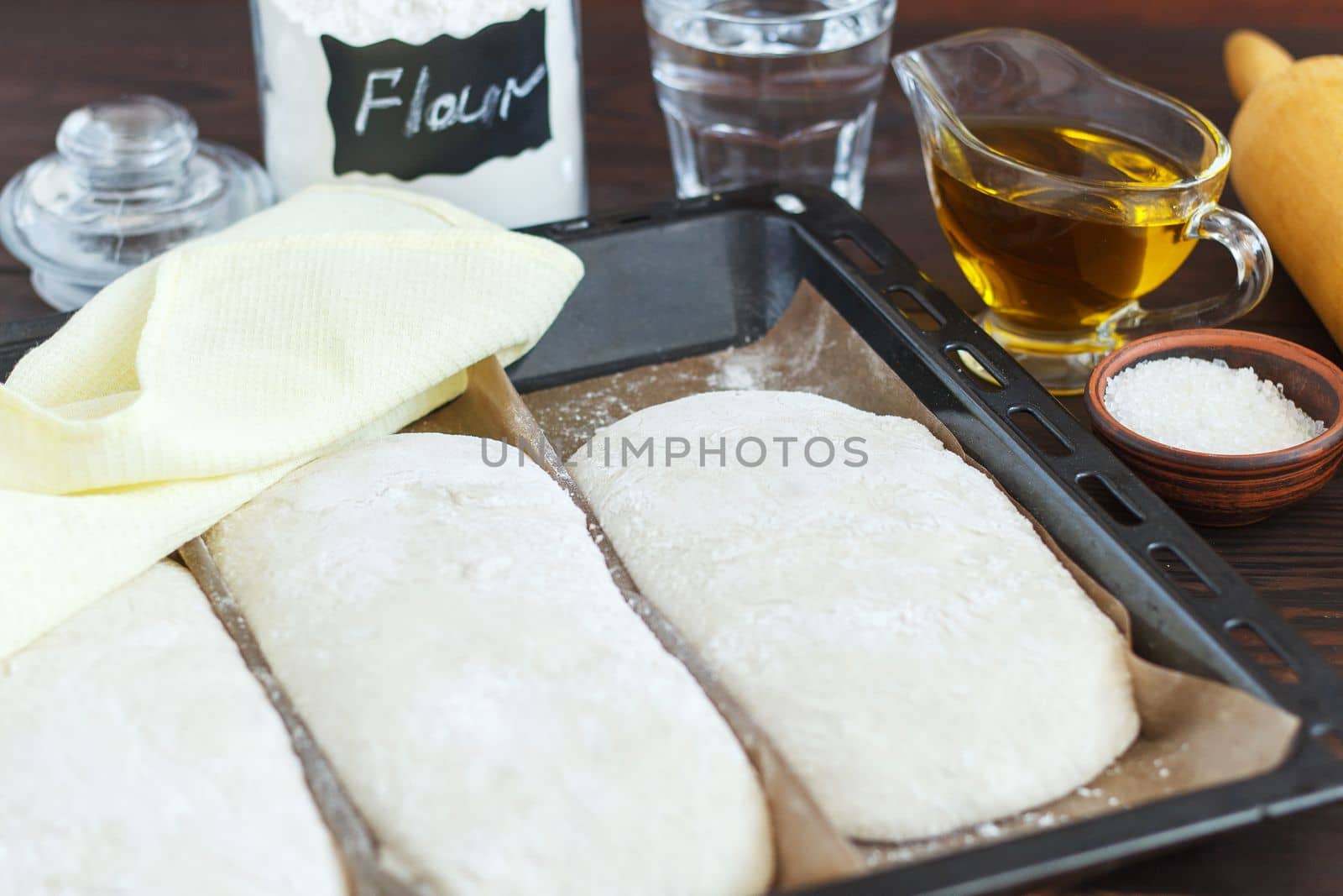 fresh raw dough for baking bread on a baking sheet, ready for baking with ingredients: flour, olive oil, water, salt on a wooden background.