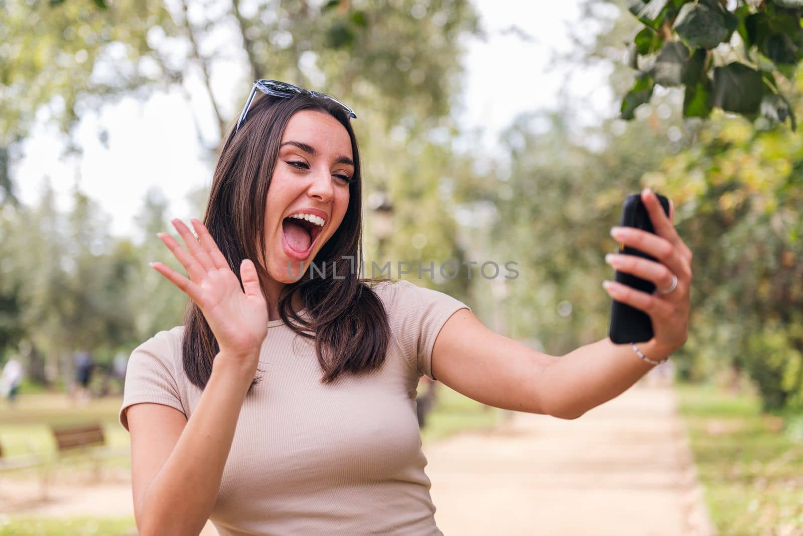 young caucasian woman waving happily during a video call with her mobile phone from a park, concept of youth and technology of communication