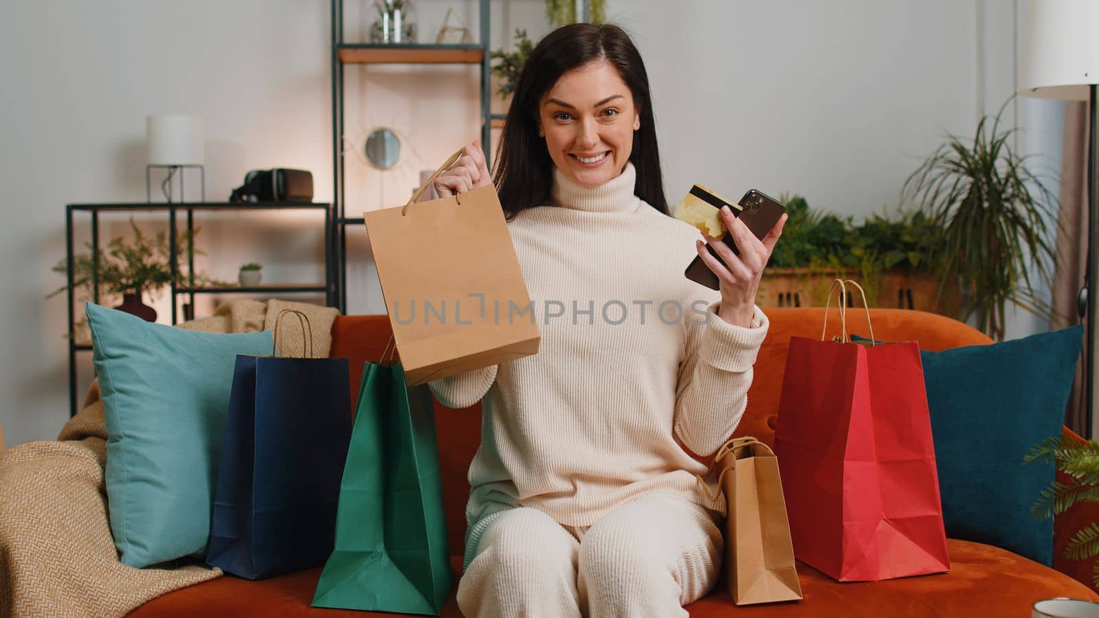 Happy shopaholic consumer young woman sitting with shopping bags at home apartment sitting on couch making online order, payment with credit plastic bank card. Discount sale on internet store mall