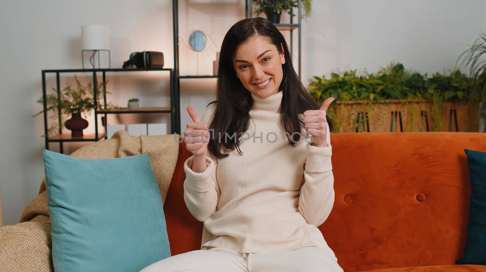 Like. Happy excited brunette woman looking approvingly at camera showing thumbs up, like sign positive something, good great news, positive feedback. Adult girl sitting on couch at home living room
