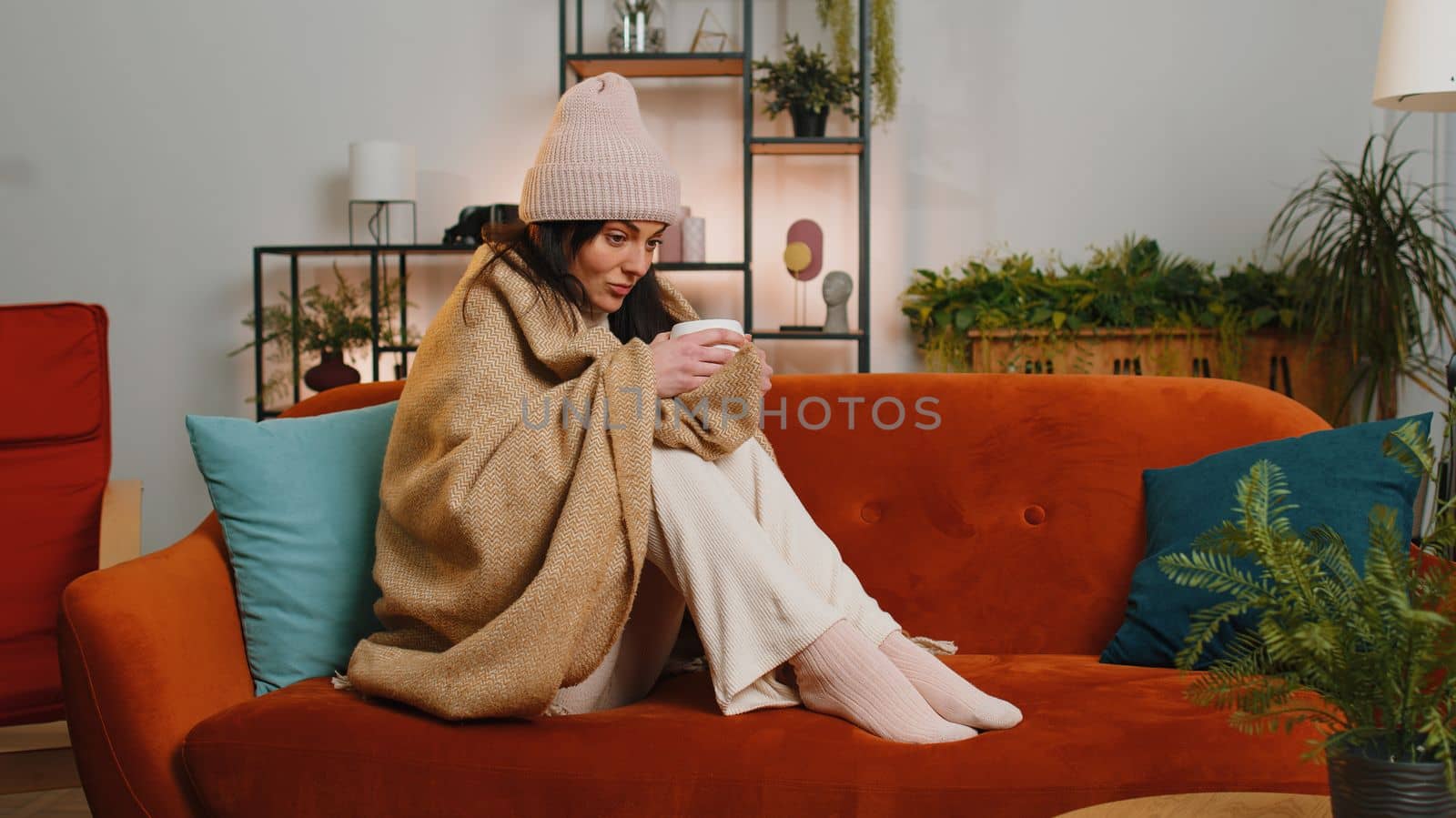Sick young woman wrapped in plaid sitting, shivering from cold on sofa drinking hot tea in unheated apartment without heating due debt. Unhealthy girl in hat feeling discomfort try to warm up at home