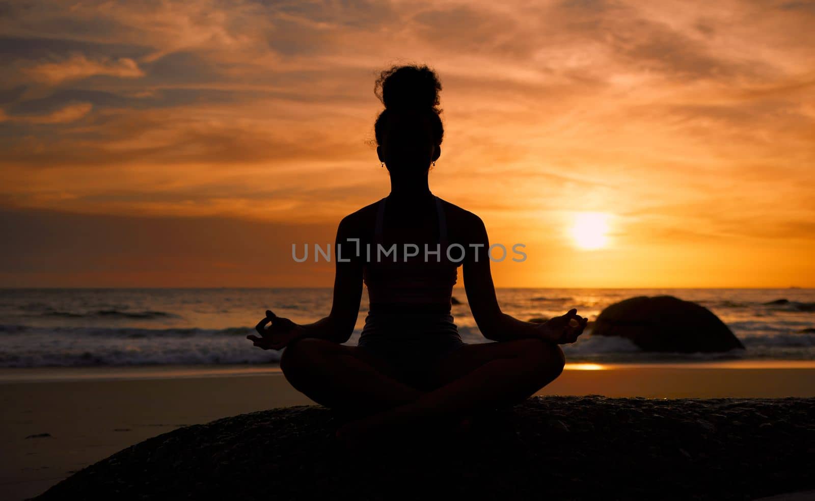 Sunset, beach and silhouette of a woman in a lotus pose while doing a yoga exercise by the sea. Peace, zen and shadow of a calm female doing meditation or pilates workout outdoor at dusk by the ocean.