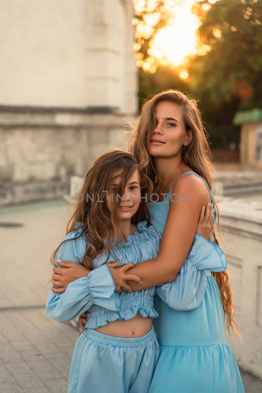 Mother aughter sunset. in blue dresses with flowing long hair against the backdrop of sunset. The woman hugs and presses the girl to her
