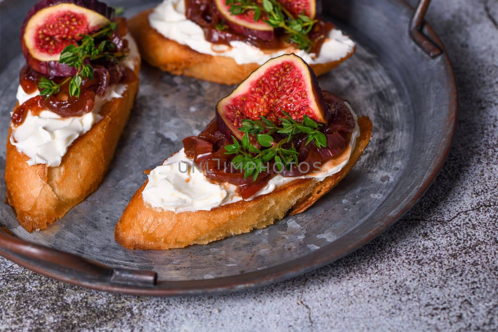 Canape or crostini with toasted baguette, cream cheese, onion jam, figs and fresh thyme on a tin tray. Ideal appetizer as an aperitif.