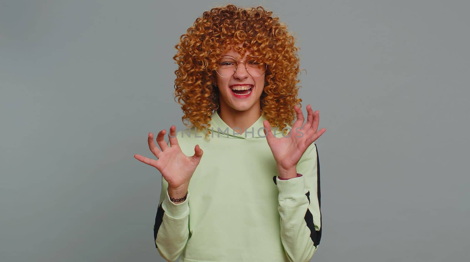 Funny furious teenager girl with curly frizzy hairstyle pretending to be a tiger, imitate parody of lion fierce shout roar, making cats claws scratching moves, flirting. Child kid on gray background