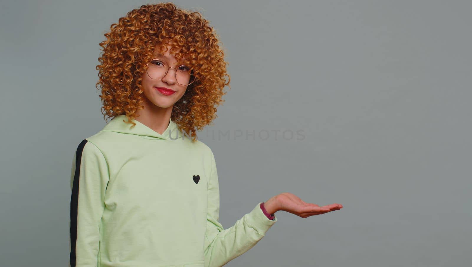 Happy teenager girl showing thumbs up pointing empty place, advertising area for commercial text, copy space for goods promotion banner, copy-space. Young child kid with curly hair on gray background