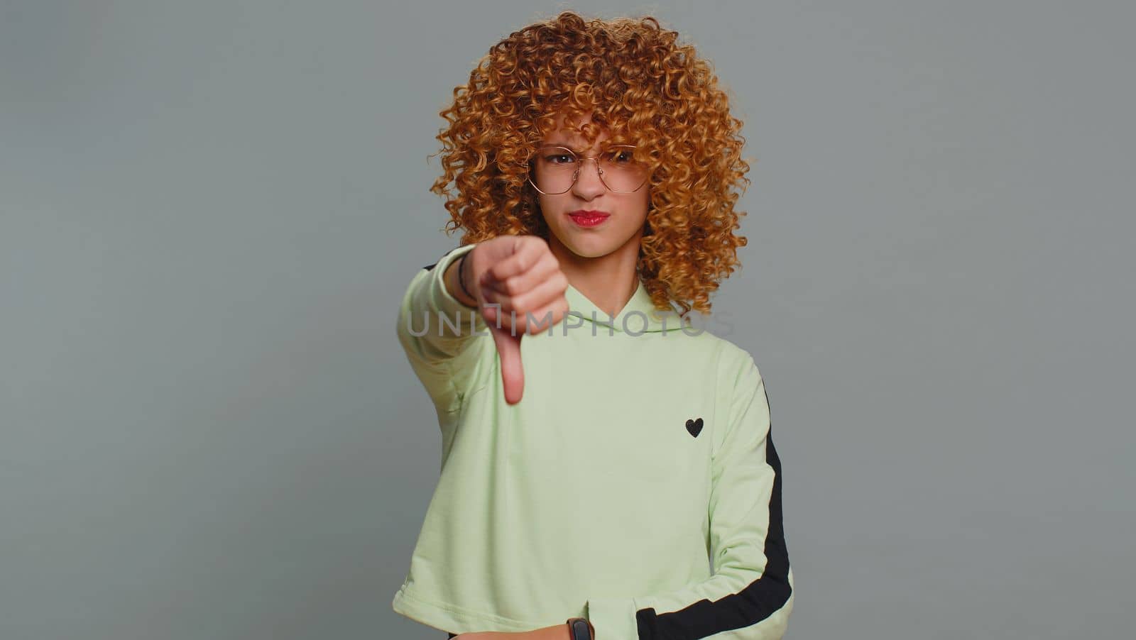 Dislike. Upset young child girl kid with curly hair showing thumbs down sign gesture, expressing discontent, disapproval, dissatisfied bad work indoors. Displeased teenager children on gray background