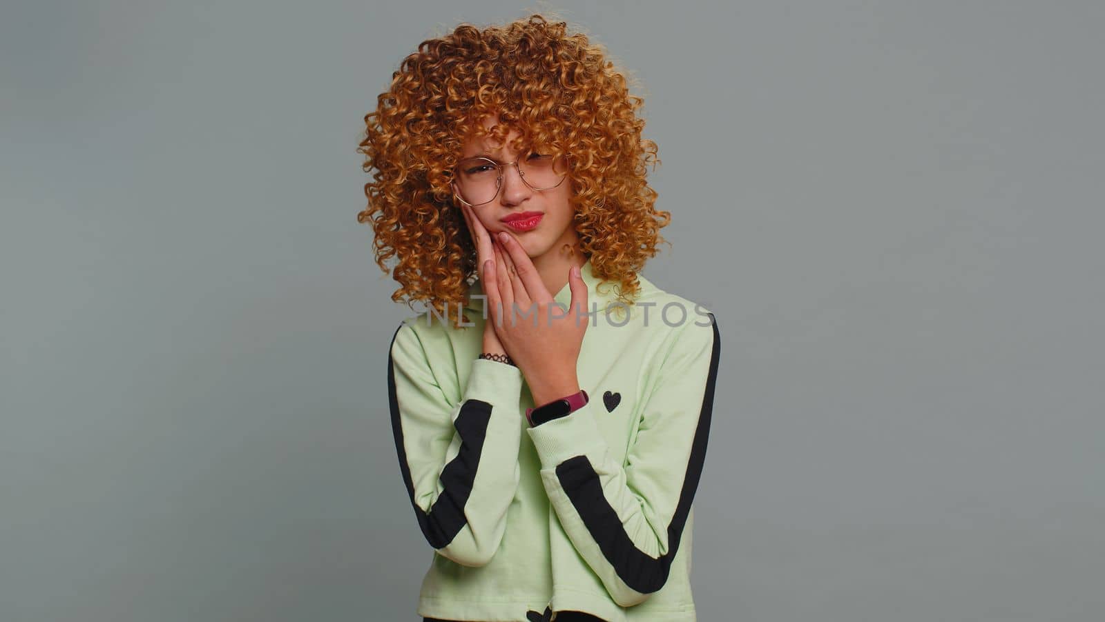 Dental problems. Young curly hair child girl kid touching cheek, closing eyes with expression of terrible suffer from painful toothache, sensitive teeth, cavities. Teenager children on gray background