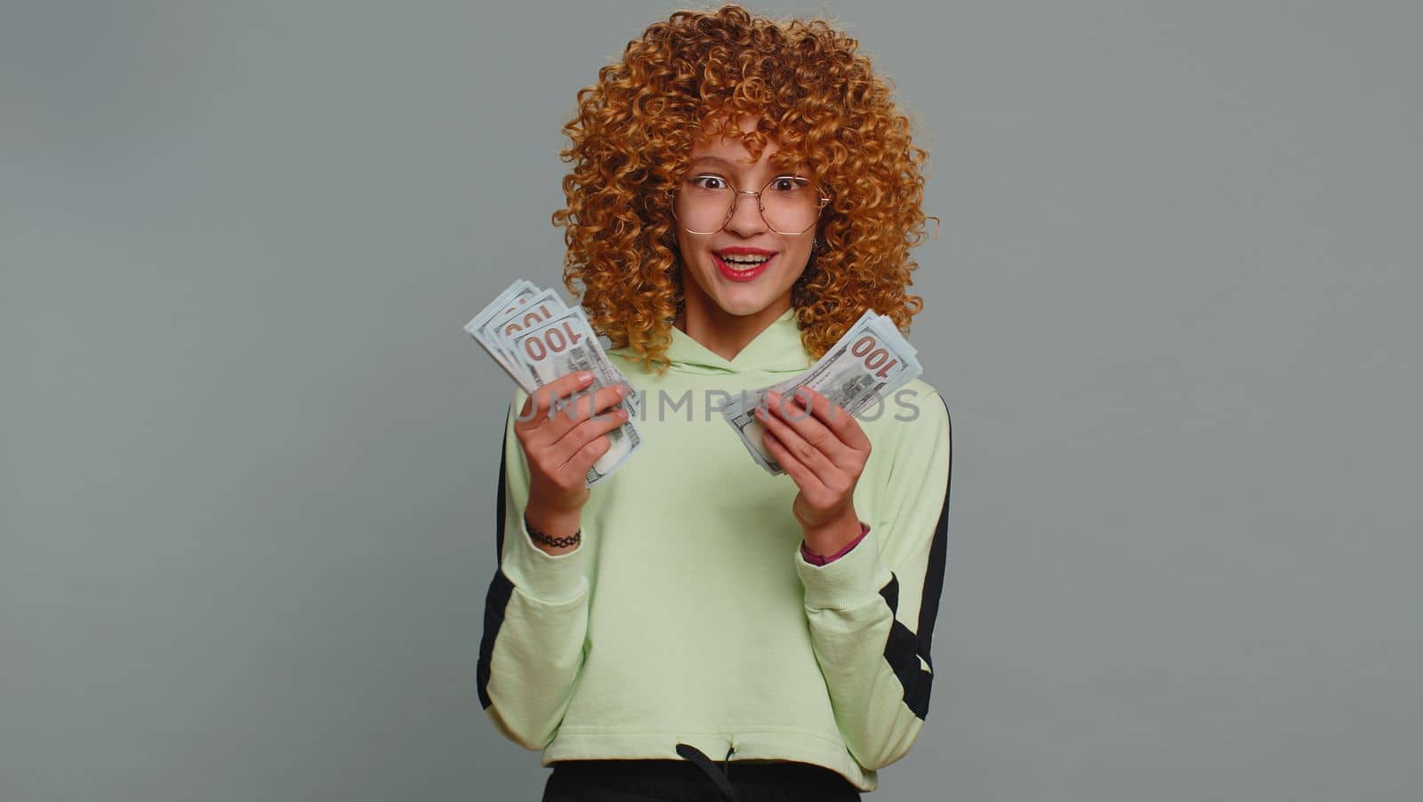 Rich pleased young teenager school girl in glasses waving money dollar cash banknotes bills like a fan, success, lottery winner, big income, wealth. Child kid with curly hair on studio gray background