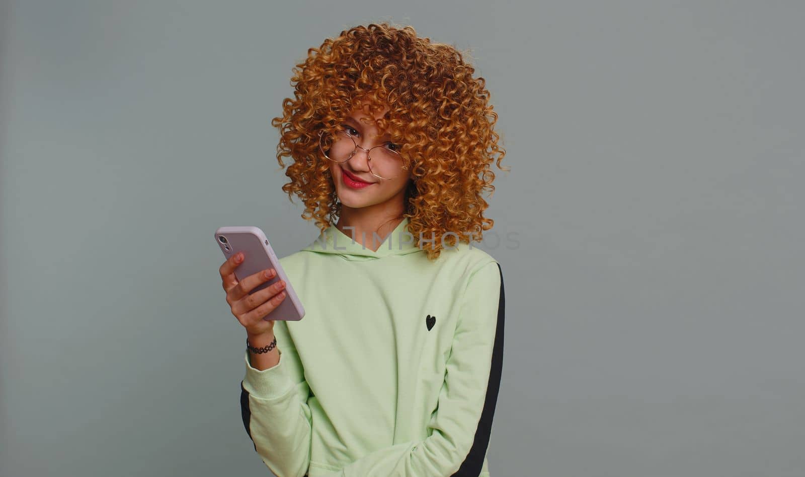 Teenager girl with curly frizzy hairstyle texting share messages content on smartphone social media applications, watching relax movie news, online store shopping sale. Child kid on gray background