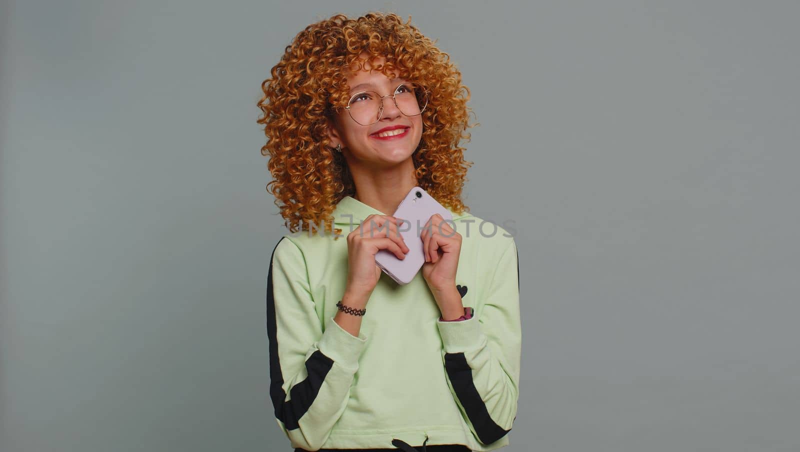 Young child girl kid with curly hair use mobile smartphone typing browsing say Wow yes found out great big win good news celebrate doing winner gesture. Teenager children on studio gray background