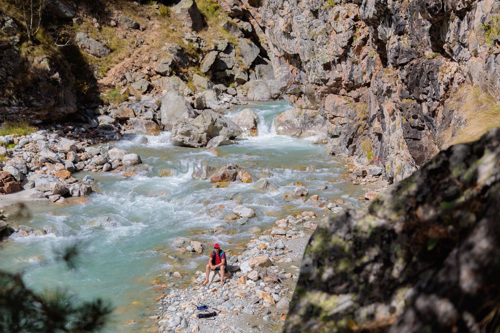 A male tourist sits by the river in the mountains. Hiking in the mountains
