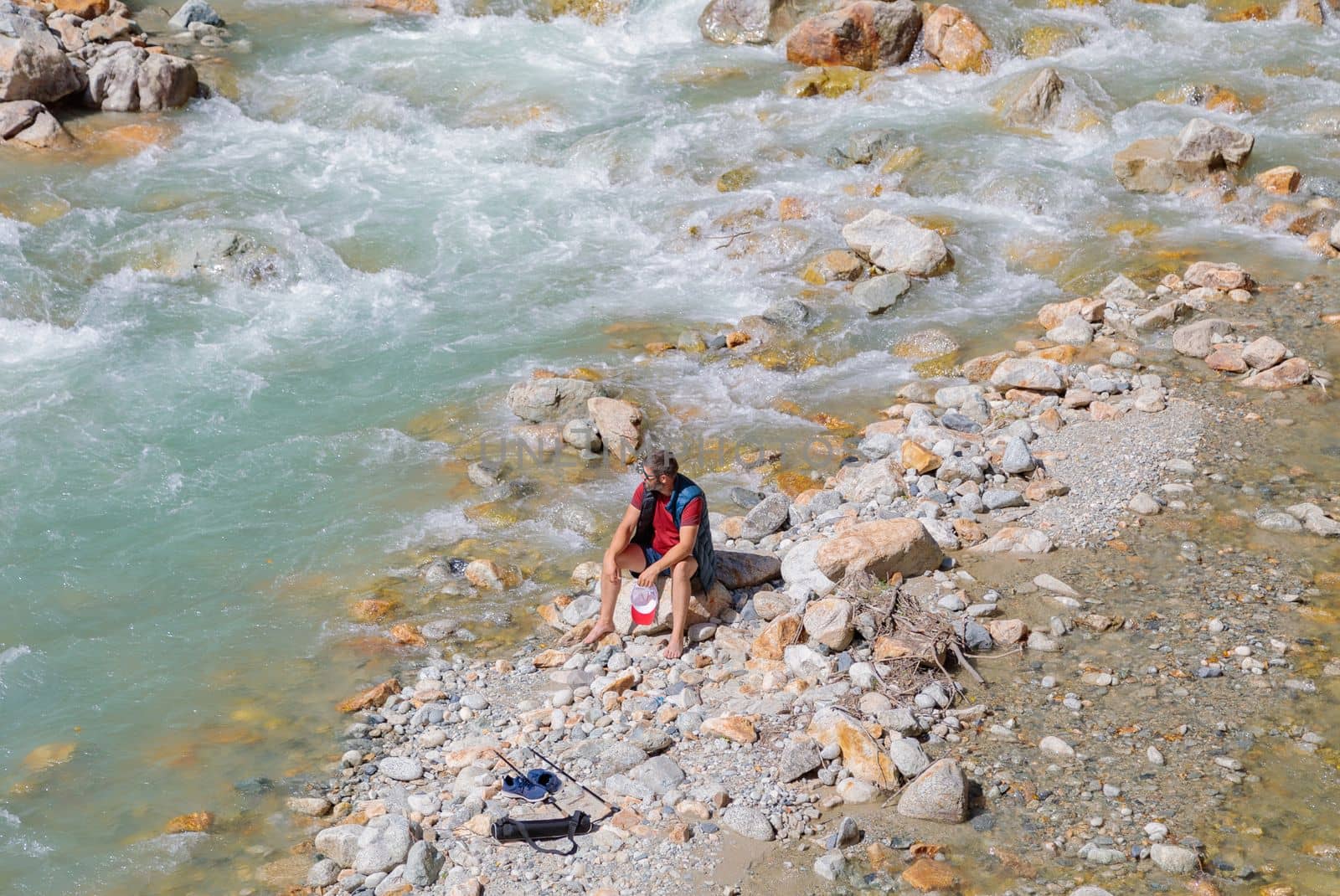 A male tourist sits by the river in the mountains. Hiking in the mountains
