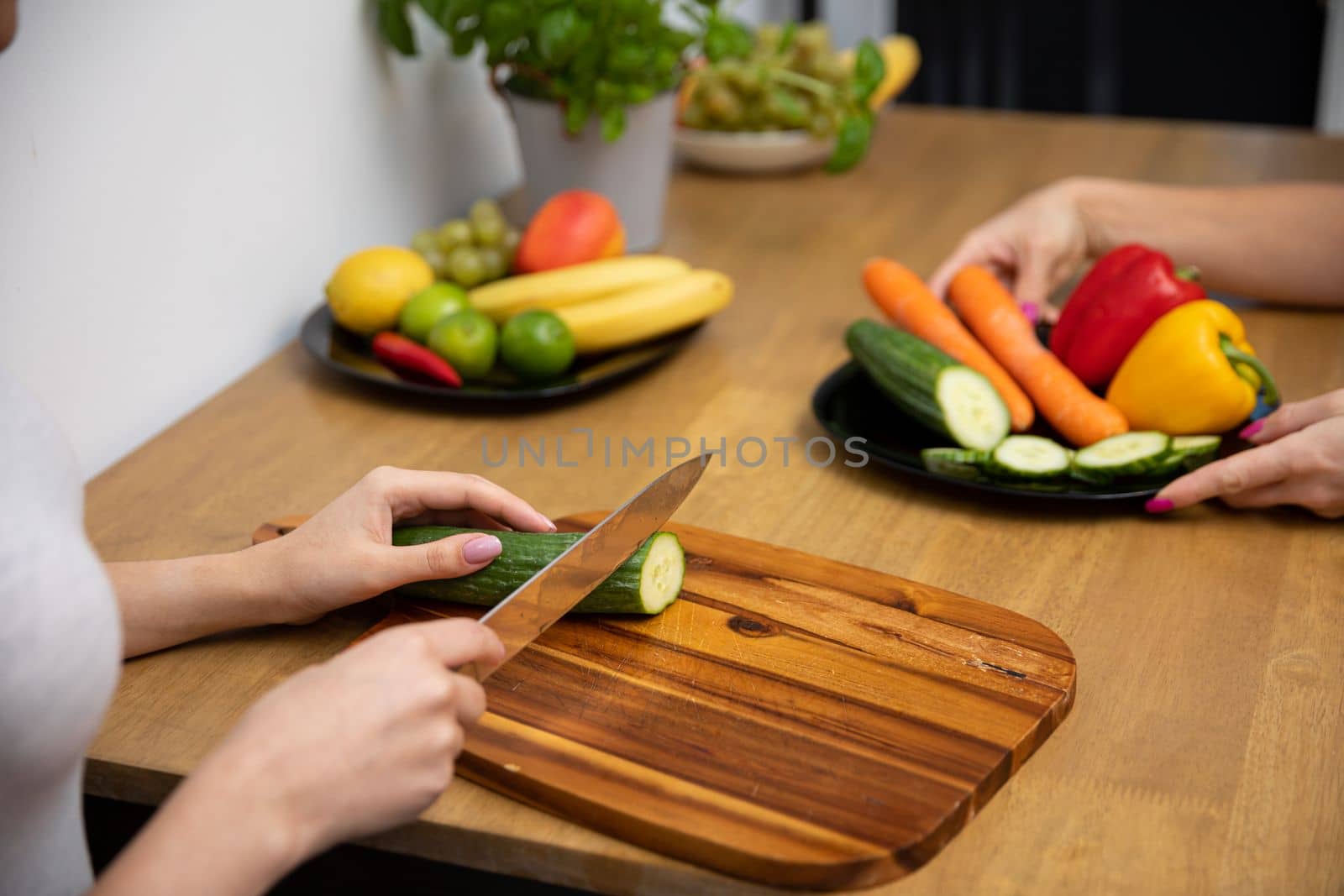 Plates of vegetables and fruits stand on a brown table. Women's hands cut a green cucumber with a knife on a brown cutting board. Preparing a healthy meal.